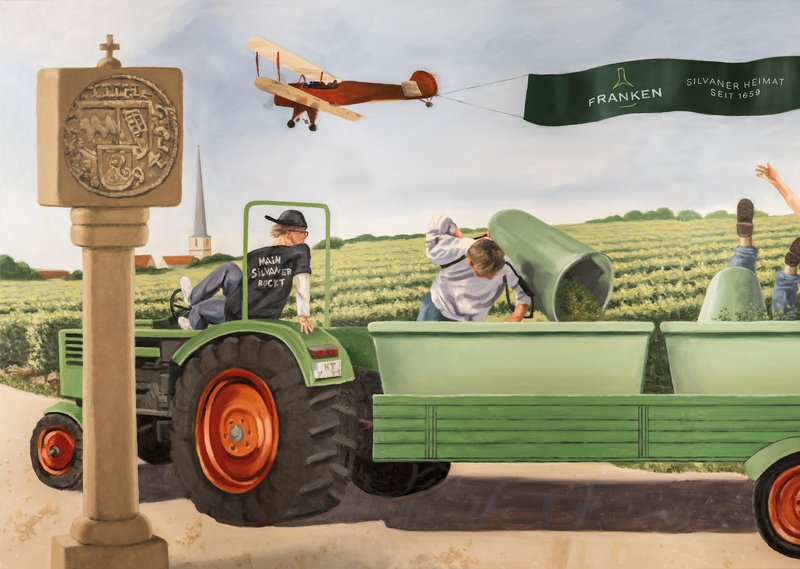 Ein junger Mann lenkt mit seinen Füßen einen Traktor an einem Weinberg vorbei. Darüber fliegt ein Doppeldecker.